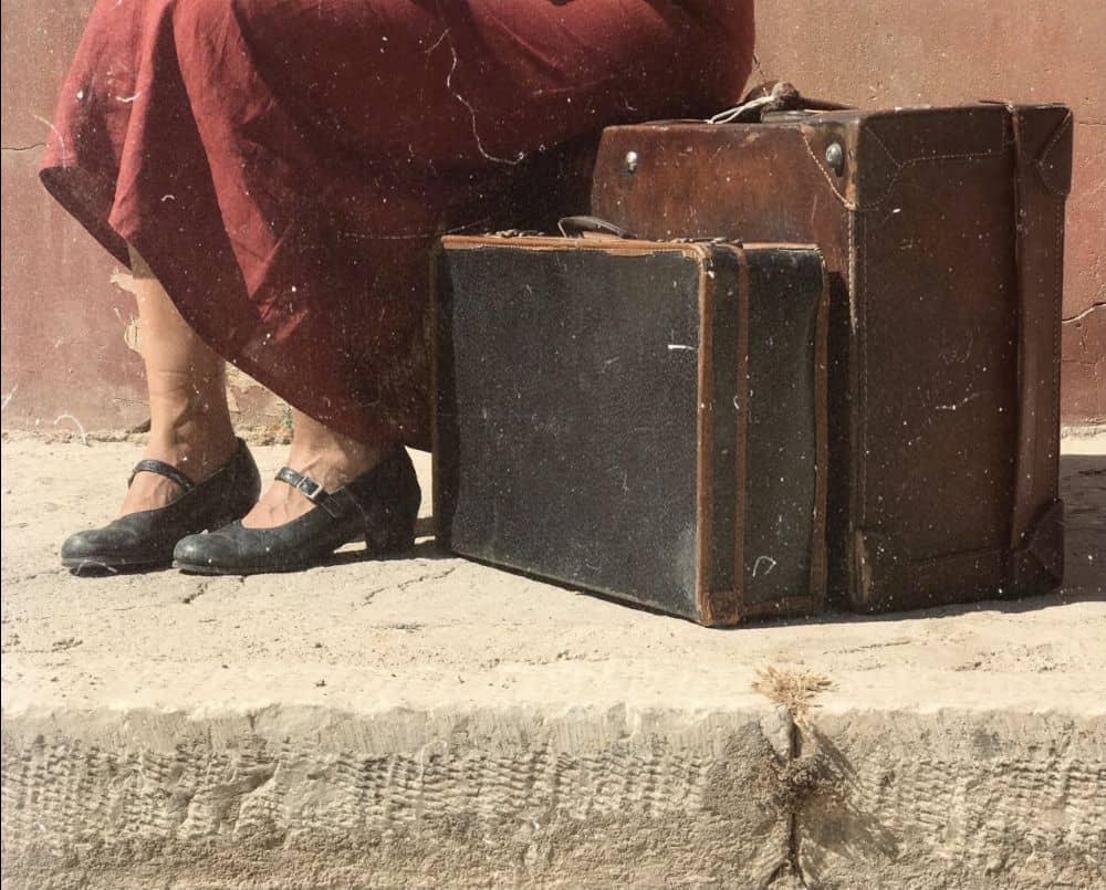Φωτογραφία εξωφύλλου για την μουσικοθεατρική παράσταση με τίτλο "Το χρονικό των δέκα ημερώ". Στην φωτογραφία απεικονίζεται ένα κόκκινο βαθύ φόρεμα μίας καθιστής γυναίκας, από τη μέση και κάτω. Καθώς, στο πλάι της υπάρχουν δύο μεγάλες βαλίτσες παλαιού τύπου. Η φωτογραφία είναι ξεφθοριασμένη μίας άλλης εποχής.