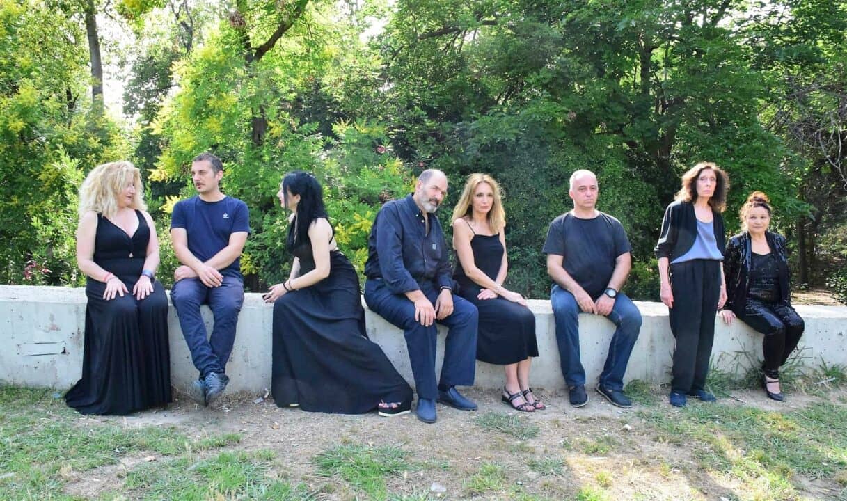 Οκτώ ηθοποιοί της παράστασης ποζάρουν για το δελτίο τύπου σε ένα πεζούλι εξωτερικού τοπίου.